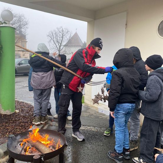 Eine Gruppe von Menschen in Winterkleidung steht um ein Feuer herum. Eine Person hält einen großen Stock mit einem Stern, wahrscheinlich zum Marshmallow-Rösten. Der Bereich ist nass, was auf kürzlichen Regen hindeutet.