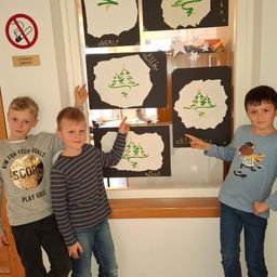 Drei Jungen stehen vor einem Fenster und zeigen auf Zeichnungen von Weihnachtsbäumen. Das Fenster hat ein Rauchverbotsschild und eine Beschriftung, die 'Lehrer-Zimmer' lautet. Die Jungen tragen Freizeitkleidung, und der Junge auf der rechten Seite trägt ein blaues T-Shirt mit einem Skateboarder darauf.