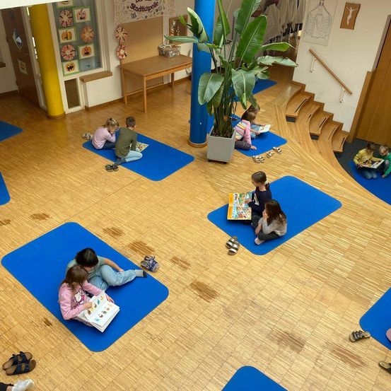 Eine Gruppe von Kindern sitzt auf blauen Matten auf einem Holzboden und liest Bücher. Eine große Topfpflanze steht in der Mitte. Links neben einer Tür befindet sich ein Tisch mit Gegenständen. Eine Treppe führt zu einem Flur.