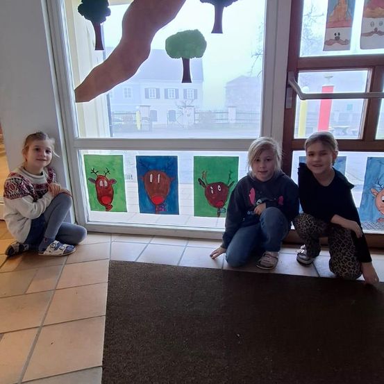 Drei junge Mädchen sitzen zusammen auf dem Boden und lächeln für ein Foto vor einem Fenster mit bunten Rentiergemälden.