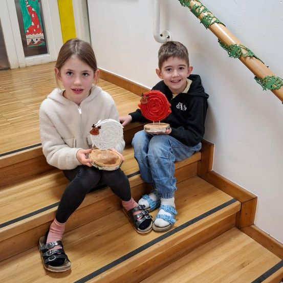 Zwei Kinder sitzen auf den Stufen und halten eine Holzbastelarbeit in den Händen. Ein Mädchen trägt eine weiße Kapuzenjacke, während ein Junge eine schwarze Kapuzenjacke trägt. Sie lächeln beide.