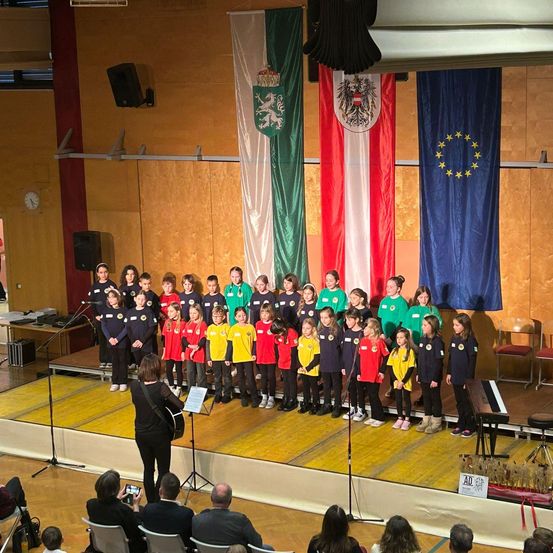 Ein Chor von Kindern in grünen, gelben und roten Uniformen steht auf der Bühne mit Flaggen dahinter. Eine Frau mit einer Gitarre steht vor ihnen, während ein Klavier in der Nähe steht. Das Publikum sitzt auf Stühlen und beobachtet die Aufführung.
