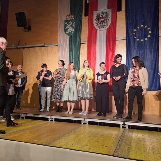 Eine Gruppe von Menschen steht auf der Bühne, klatscht und lächelt. Hinter ihnen sind Flaggen von Österreich und der Europäischen Union zu sehen.