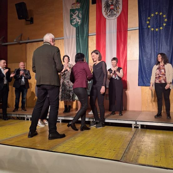 Eine Gruppe von Menschen steht auf der Bühne und klatscht in die Hände. Vier Frauen stehen vorne und lächeln. Dahinter befinden sich Flaggen Österreichs und der Europäischen Union.