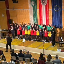 Ein Schulchor tritt auf der Bühne auf, mit Fahnen dahinter, darunter die Flagge Österreichs. Ein Publikum beobachtet sie von Stühlen aus.