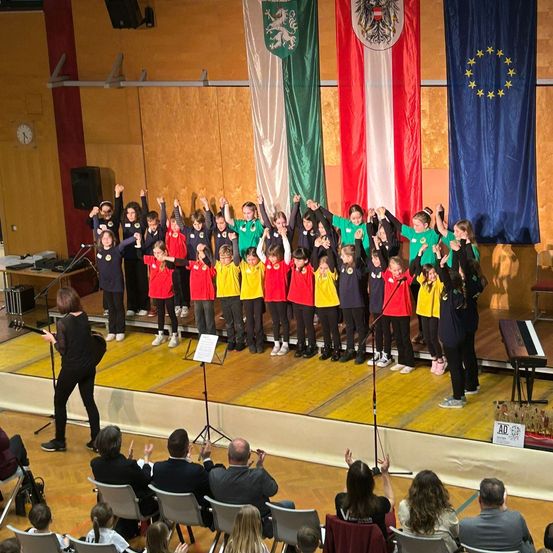 Ein Schulchor tritt auf der Bühne auf, mit Fahnen dahinter, darunter die Flagge Österreichs. Ein Publikum beobachtet sie von Stühlen aus.