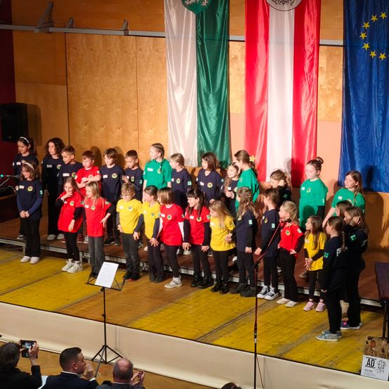 Eine Gruppe von Kindern in roten und grünen Uniformen steht auf der Bühne, wahrscheinlich als Chor. Sie halten Mikrofone und hinter ihnen stehen Flaggen verschiedener Länder. Die Bühne hat Holzboden. Das Publikum schaut zu.