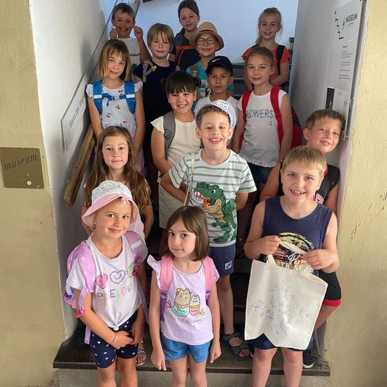 Eine Gruppe von Kindern, wahrscheinlich Schüler, steht auf einer Treppe, lächelt und posiert für ein Foto. Sie sind lässig gekleidet, einige mit Rucksäcken, und scheinen sich in einem Museum zu befinden.