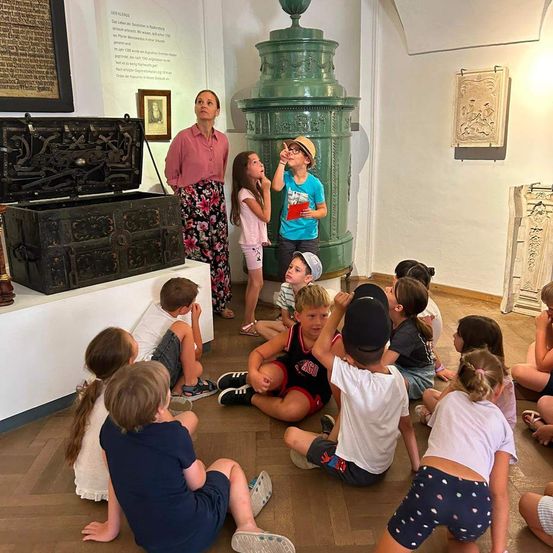 Eine Gruppe von Kindern sitzt auf dem Boden in einem Museum und hört einer Frau zu, die in der Nähe eines großen grünen Objekts steht. Die Kinder schauen zu der Frau und dem Objekt auf.