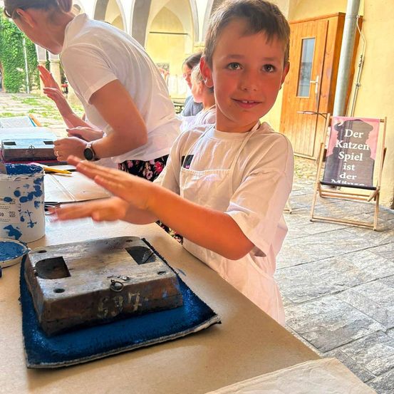 Ein junger Junge in einer weißen Schürze lächelt, während er an einem Projekt an einem Tisch arbeitet, mit einem hölzernen Objekt und Farbdosen in der Nähe.