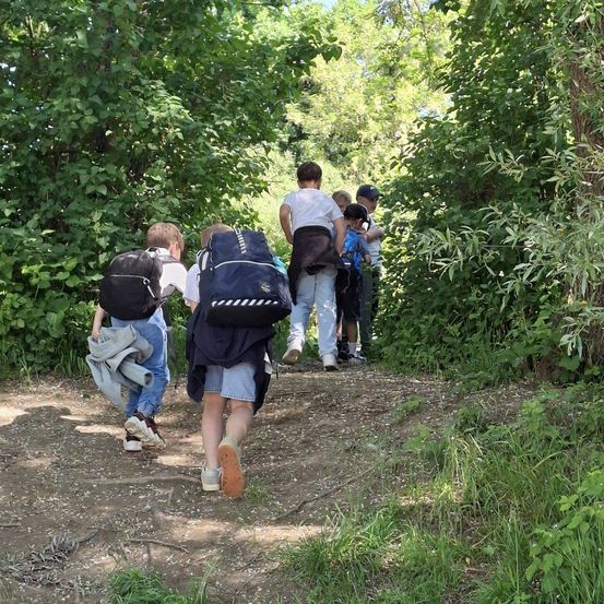 Eine Gruppe von Kindern, die auf einem Pfad im Wald laufen, einige tragen Rucksäcke und Jacken.