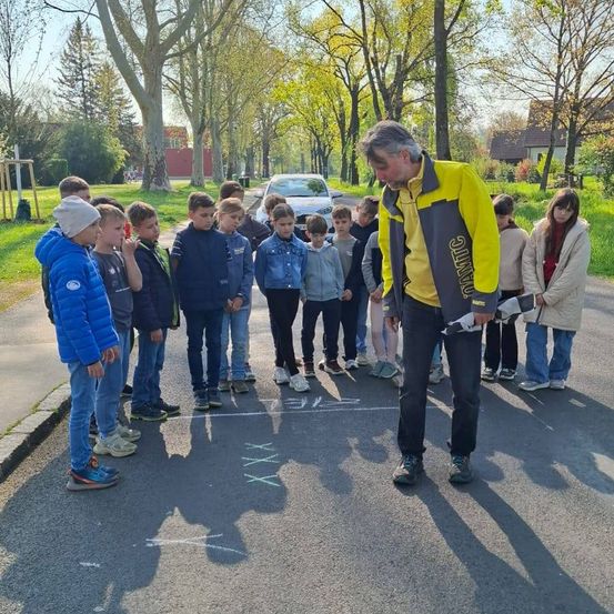 Eine Gruppe von Kindern und ein erwachsener Mann stehen auf einer Straße mit Kreidezeichnungen, möglicherweise an einem Spiel beteiligt. Der Mann trägt eine gelbe Jacke und hält ein Stück Papier.