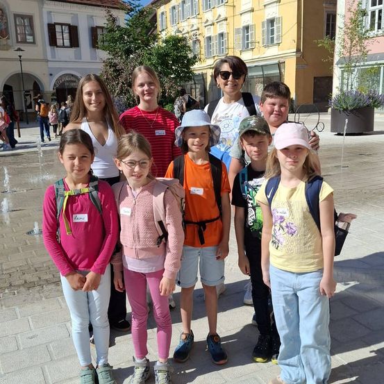 Eine Gruppe von Kindern und einer erwachsenen Frau stehen auf einem Platz und lächeln für ein Foto. Sie tragen Freizeitkleidung und einige haben Rucksäcke. Hinter ihnen stehen Gebäude, ein Brunnen und gehende Menschen.