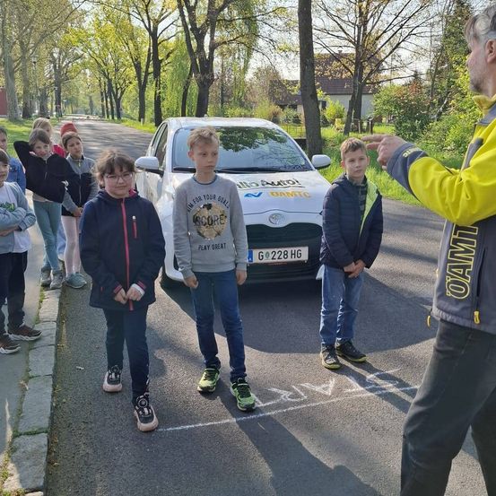 Eine Gruppe von Kindern und ein Mann in einer gelben Jacke stehen neben einem weißen Auto auf der Straße, wobei der Mann auf das Auto zeigt.