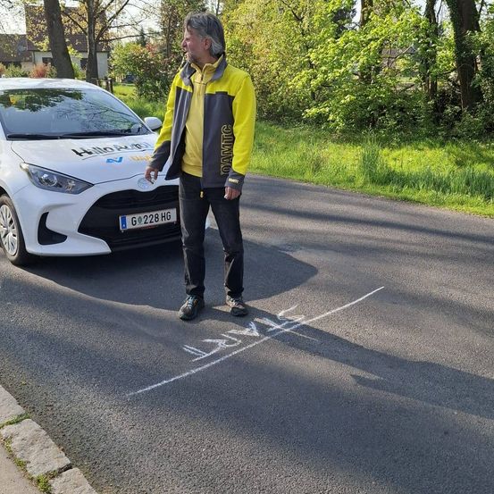 Ein Mann in einer gelben und grauen Jacke steht neben einem weißen Toyota Yaris mit dem Kennzeichen G 2228 HG. Er schaut auf die Straße, wo weiße Kreidemarkierungen sichtbar sind.