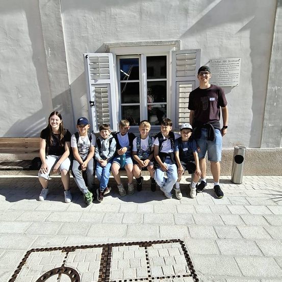 Eine Gruppe von Kindern und zwei Erwachsenen posiert für ein Foto vor einem Gebäude mit weißen Fensterläden und einer Plakette an der Wand.