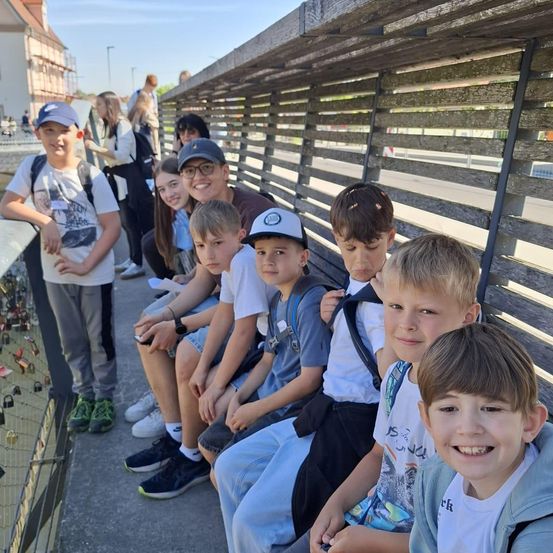 Eine Gruppe von Kindern und einem Mann sitzen auf einer Brücke mit einem Holzgeländer, einige tragen Hüte und Brillen. Sie lächeln und posieren für ein Foto. Hinter ihnen befinden sich Gebäude, Bäume und ein klarer Himmel.