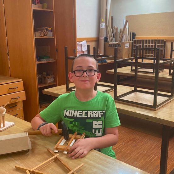 Ein junger Junge mit einer grünen Minecraft-T-Shirt und einer Brille arbeitet in einer Werkstatt an einem Holzprojekt an einem Tisch.