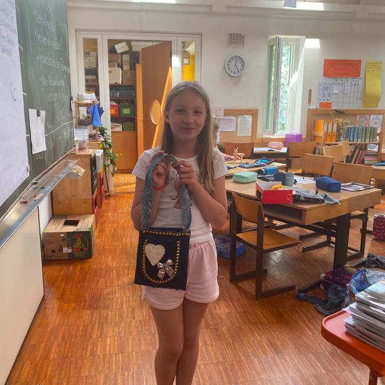 Ein junges Mädchen in einem Klassenzimmer hält eine kleine Tasche mit Herzdesign. Der Klassenraum ist mit Tischen, Stühlen und einer Tafel gefüllt.