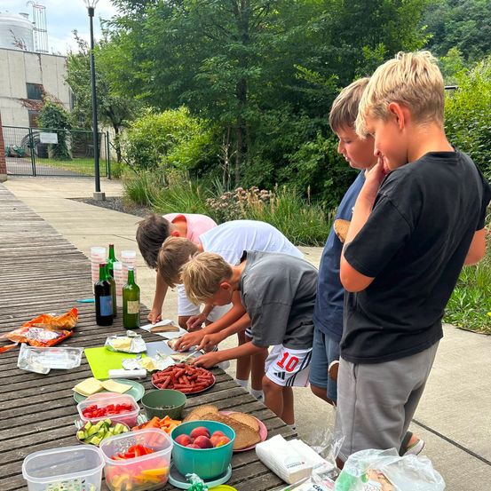 Vier Jungen bereiten draußen auf einem Holztisch Essen zu. Sie haben verschiedene Zutaten und Utensilien vor sich ausgebreitet. Dahinter steht ein Gebäude mit einem Zaun und Bäumen.