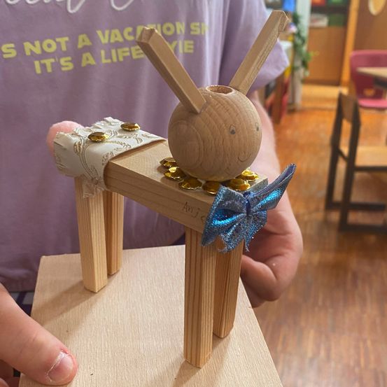 Eine Person hält eine Holzfigur mit einer blauen Schleife und goldenen Münzen. Die Figur hat einen runden Kopf und Ohren. Sie steht auf einer Holzbasis.