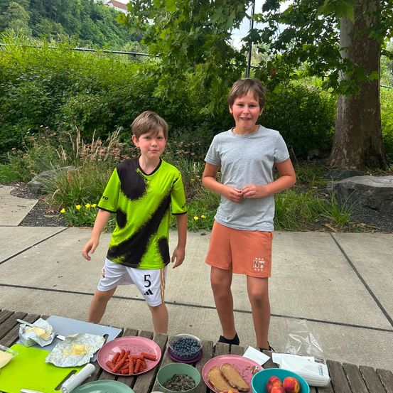 Zwei junge Jungen stehen neben einem Picknicktisch mit Essen darauf. Ein Junge trägt ein grünes und schwarzes Fußballtrikot mit der Nummer 5. Der andere Junge trägt ein graues T-Shirt und orange Shorts.