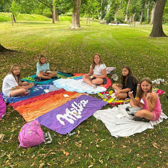 Eine Gruppe junger Mädchen sitzt auf Decken in einem Park, einige essen Snacks. Die Decken sind bunt, und auf einer steht das Wort 'Mila'. Die Mädchen sind von Bäumen und Gras umgeben.