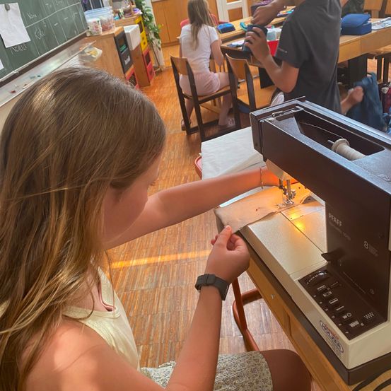 Ein junges Mädchen sitzt an einer Nähmaschine, trägt eine Uhr und ein Armband. Hinter ihr sitzt ein Junge an einem Schreibtisch und hält einen Becher. Im Hintergrund sitzt ein weiteres Mädchen an einem Tisch.