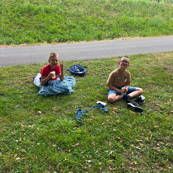 Zwei Jungen sitzen auf dem Gras, einer mit einem Friedenszeichen und einem Becher, der andere mit einem Rucksack und einem Schuh in der Nähe.