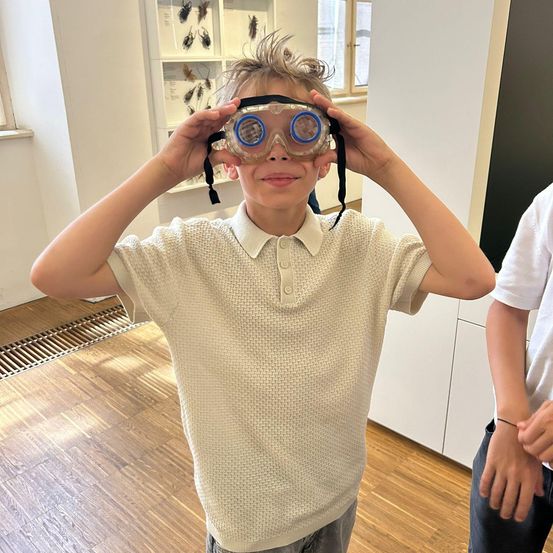 Ein junger Junge mit einer Brille auf der Stirn, lächelnd, in einem Raum mit Holzböden und weißen Wänden.