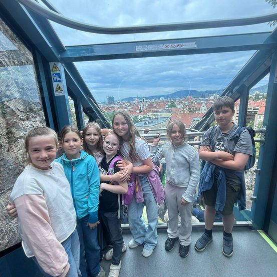 Eine Gruppe junger Kinder, sowohl Jungen als auch Mädchen, steht in einer glasverschlossenen Kabine und posiert für ein Foto. Sie sind auf einer Seilbahnfahrt mit Blick auf eine Stadt unten.