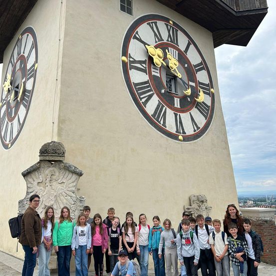 Eine Gruppe von Kindern und Erwachsenen steht vor einem großen Uhrturm mit zwei Uhren, eine zeigt die Zeit 10:30 an.