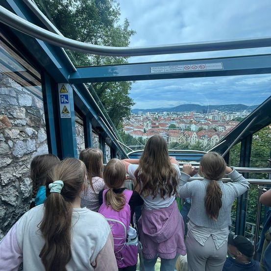 Eine Gruppe von Kindern in einer Seilbahn, die eine Stadtlandschaft mit Gebäuden und Bäumen unter einem bewölkten Himmel betrachten.