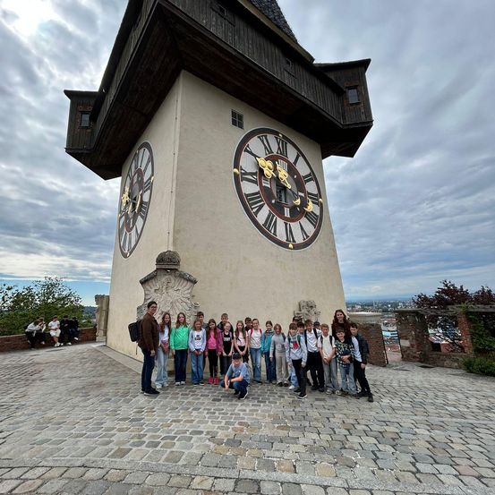 Eine Gruppe von Kindern und zwei Erwachsenen posiert vor einem großen Uhrenturm mit zwei Uhren auf einem Kopfsteinpflasterplatz.