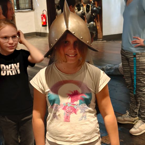 Ein junges Mädchen mit einem mittelalterlichen Helm lächelt in die Kamera, mit einem Jungen mit Brille und einem Mädchen in Freizeitkleidung im Hintergrund.