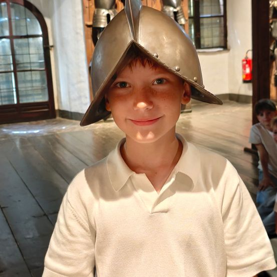 Ein junger Junge mit einem mittelalterlichen Helm lächelt in die Kamera in einem Museum. Hinter ihm ist ein Feuerlöscher an der Wand angebracht.