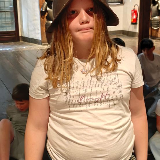 Ein junges Mädchen mit blonden Haaren trägt einen braunen Hut und ein weißes T-Shirt mit rosa Text und steht in einem Raum mit Holzböden und einem an der Wand montierten Feuerlöscher.