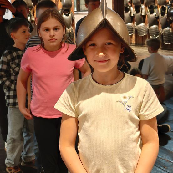 Ein junges Mädchen mit einem mittelalterlichen Helm steht vor einer Gruppe von Kindern, von denen einige Rüstungen tragen. Der Schauplatz scheint ein historisches Ausstellungs- oder Museumsgebäude zu sein.
