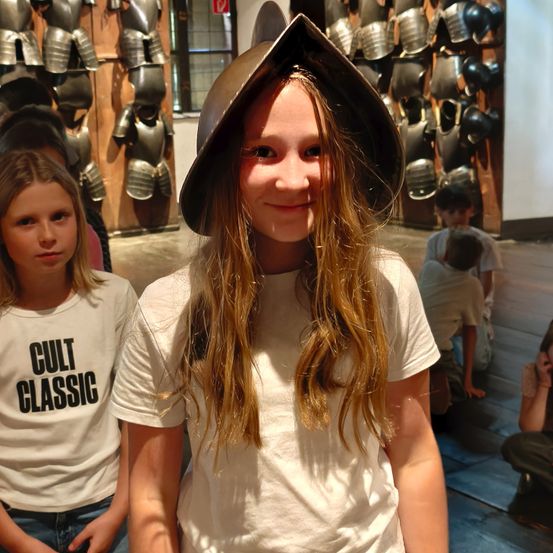 Ein Mädchen in einem mittelalterlichen Hut lächelt in einem Museum, mit Rüstungen an der Wand und einem anderen Mädchen in einem weißen Shirt mit 'Cult Classic' darauf.
