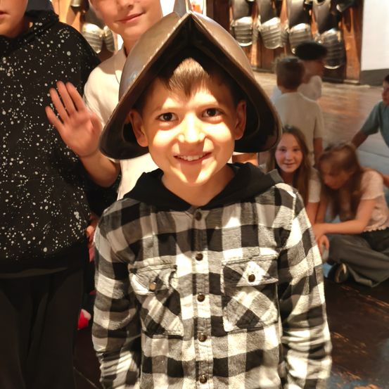 Ein junger Junge mit einem Metallhelm und einem schwarz-weiß karierten Hemd lächelt in die Kamera. Hinter ihm sitzen mehrere Kinder auf dem Boden, und einige stehen. Der Hintergrund ist unscharf mit ausgestellten Rüstungsteilen.