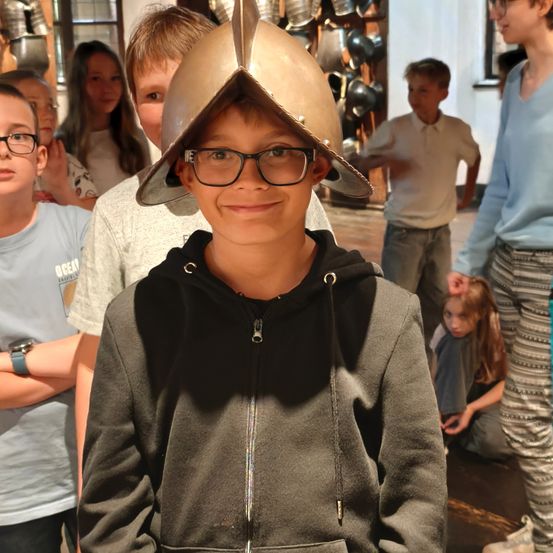 Ein junger Junge, der einen mittelalterlichen Helm trägt, lächelt für die Kamera, während mehrere Kinder und Erwachsene in einem Raum mit mittelalterlichen Artefakten hinter ihm stehen.