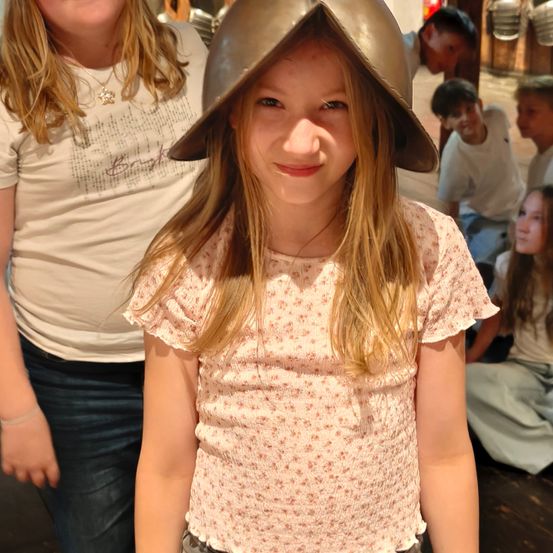 Ein junges Mädchen mit einem glänzenden Metallhelm lächelt in die Kamera. Hinter ihr steht eine Frau in einem weißen T-Shirt und Jeans, und mehrere Kinder sitzen im Hintergrund.