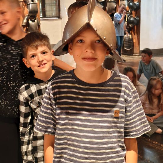 Ein Junge mit einem mittelalterlichen Helm steht neben einem anderen Jungen in einem karierten Shirt, beide lächeln in einer Museumsumgebung.