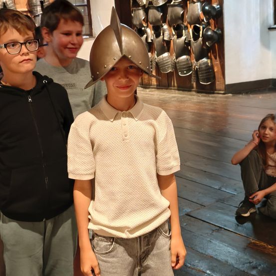 Drei Jungen stehen in einem Raum mit Holzböden und einer Wand voller mittelalterlicher Rüstungen. Ein Junge trägt einen silbernen Helm.