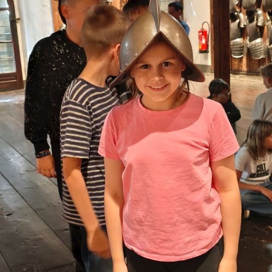 Ein junges Mädchen mit einem silbernen Helm lächelt in einem Museum, mit einem Jungen in einem gestreiften T-Shirt hinter ihr und einem Mann in einem schwarzen T-Shirt neben ihm.