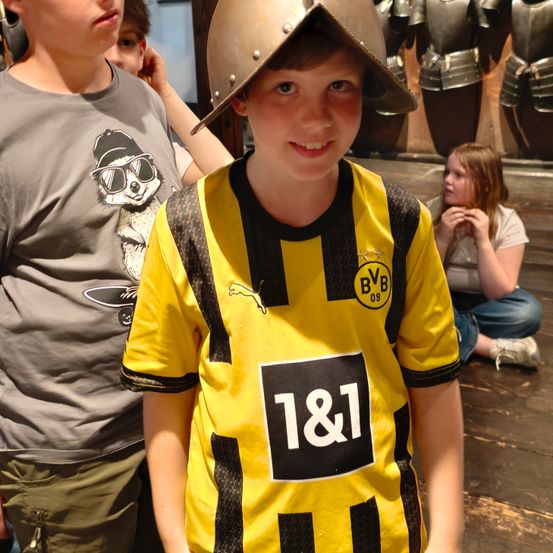 Ein Junge in einem gelben Fußballtrikot mit der Nummer 1&1 darauf, vor einer Wand mit Rüstungsmannequins stehend.