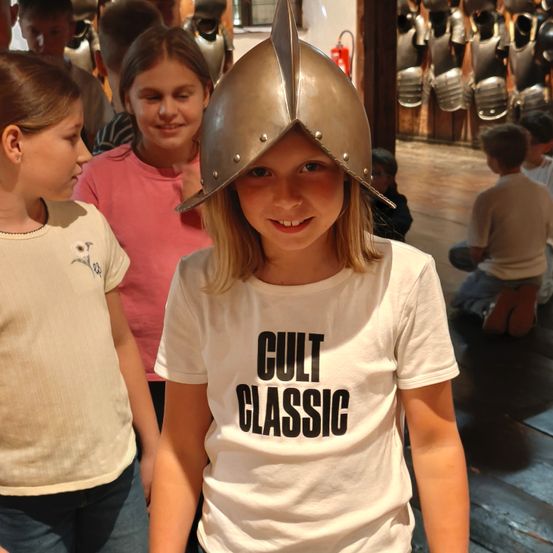 Ein junges Mädchen mit einem silbernen Helm und einem weißen T-Shirt mit den Worten 'Cult Classic' steht in einem Museum mit anderen Kindern und Rüstungen im Schaufenster.