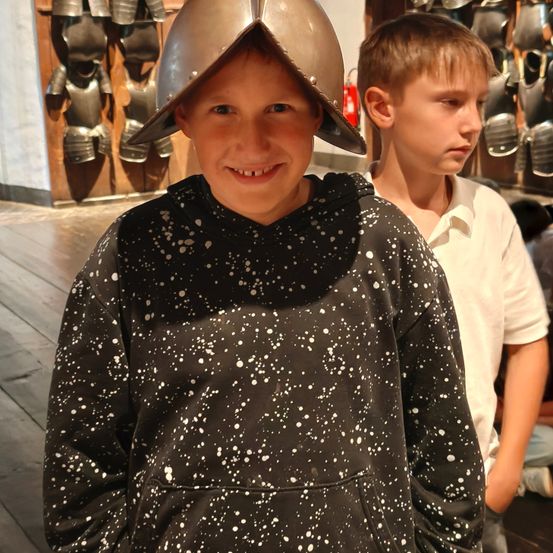Ein junger Junge mit einem silbernen Helm mit weißen Punkten auf einem schwarzen Hoodie steht neben einem anderen Jungen in einem weißen Hemd in einem Museum.