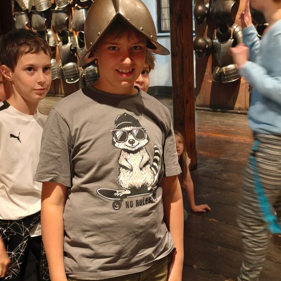 Ein Junge mit einem mittelalterlichen Helm und einem Waschbär-T-Shirt steht in einem Museum zwischen anderen Kindern.