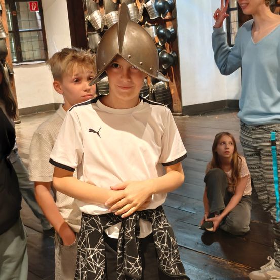 Ein junger Junge mit einem mittelalterlichen Helm steht zwischen zwei anderen Kindern in einem Raum mit Holzböden und einer mit Rüstungen verzierten Wand.